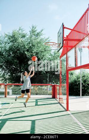 Vista laterale del corpo della giovane donna sportiva che esegue il salto mentre gioca a basket da sola sul campo sportivo Foto Stock