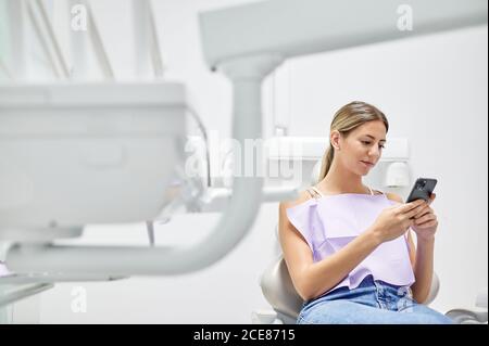 Paziente femminile con tovagliolo seduto sulla sedia e smartphone di navigazione durante l'appuntamento in clinica dentale contemporanea Foto Stock