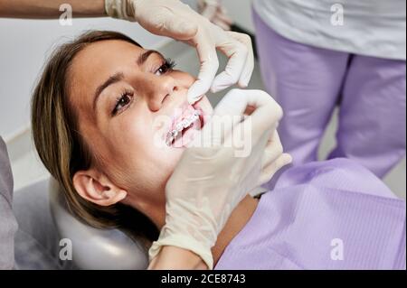Dall'alto del medico dentistico senza volto che controlla i denti di femmina il cliente indossa le bretelle mentre usa lo specchio per bocca Foto Stock