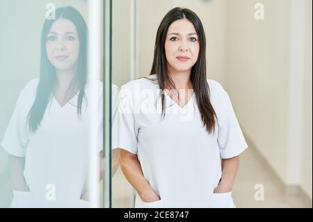 Una donna sorridente e professionale con stomatologa che indossa una divisa grigia con mano in mano la tasca che guarda la fotocamera in ospedale Foto Stock