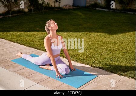 Vista laterale della donna serena seduta sul tappetino in Ardha Kapotasana a bordo piscina e meditando con gli occhi chiusi mentre si pratica yoga in cortile in estate Foto Stock