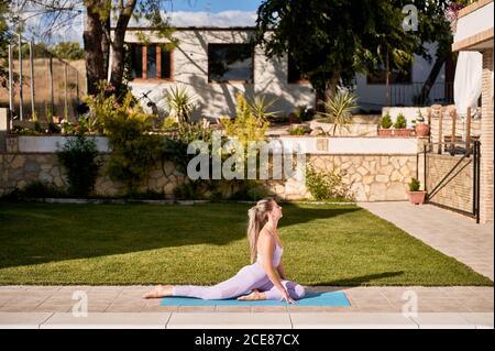 Vista laterale della donna serena seduta sul tappetino in Ardha Kapotasana a bordo piscina e meditando con gli occhi chiusi mentre si pratica yoga in cortile in estate Foto Stock