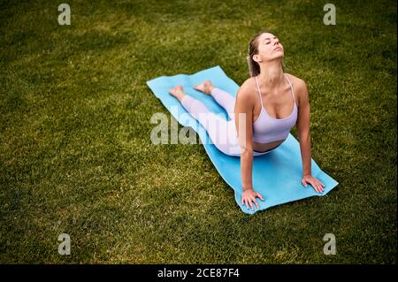 Vista dall'alto laterale di una tranquilla donna che pratica yoga Tappetino in Bhujangasana con occhi chiusi sul prato in cortile Foto Stock