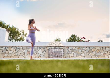 Donna tranquilla in leggings sportivi e reggiseno equilibrante sulla gamba In Vrksasana con gesto Namaste mentre si fa yoga e guardare via Foto Stock