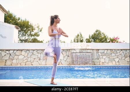 Donna tranquilla in leggings sportivi e reggiseno equilibrante sulla gamba In Vrksasana con gesto Namaste mentre si fa yoga e guardare via Foto Stock