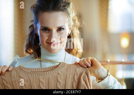 sorridente donna trendy di 40 anni in maglione bianco e gonna nel moderno soggiorno in sole giornate invernali che tiene maglioni su appendiabiti di rame. Foto Stock