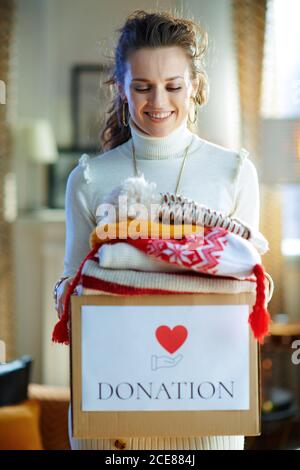 casalinga trendy di mezza età in maglione bianco e gonna con scatola di donazione con vecchi abiti caldi a casa moderna in sole giorno d'inverno. Foto Stock