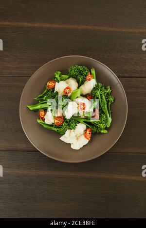 Vista dall'alto della piastra rotonda con broccolini verdi freschi e. formaggio bianco con pomodori secchi tritati su tavola di legno Foto Stock