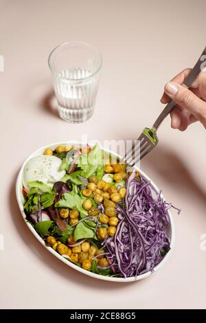 Crop persona mangiare insalata con insalata mista e cavolo rosso con ceci mentre si ha un bicchiere d'acqua Foto Stock