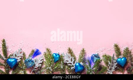 Ornamenti natalizi a forma di cuore blu con luci di festa a LED blu su sfondo rosa pastello, decorati con rami di abete rosso. Bordo inferiore. Foto Stock