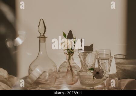 Alto angolo di fiori bianchi delicati in vaso piccolo messo su tavolo coperto di tulle e arrangiato con stile vintage vetri e decanter Foto Stock