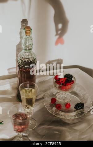 Dall'alto bottiglia di vino rosato fatto in casa versato in cristallo bicchiere vicino alla tromba di champagne e servito con piatto di fresco lamponi e more contro parete bianca con ombra a mano Foto Stock