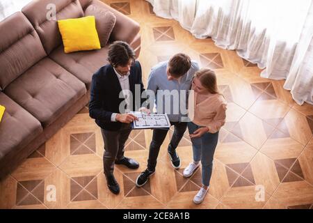 Realtor che mostra il piano della casa a coppie di giovani clienti al chiuso, vista dall'alto Foto Stock