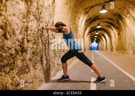 Vista laterale di una atleta anonima in vestiario sportivo e la fascia da braccio è appoggiata contro una parete ruvida mentre si allungano le gambe durante il riscaldamento in galleria con luce artificiale Foto Stock