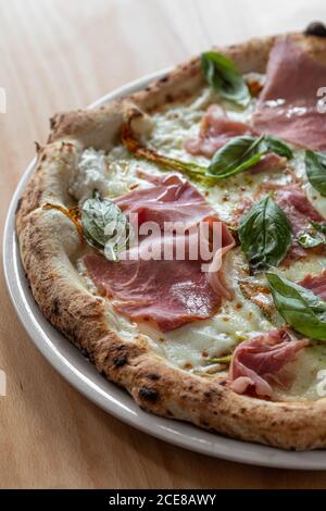 Un angolo di pizza italiana deliziosa con pomodori piccoli e. cornicione di charme servito con prosciutto e mozzarella e decorato con foglie di basilico poste su un piatto nero su un tavolo di legno Foto Stock