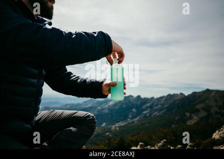 Vista laterale di un uomo dal bearded tagliato e non riconoscibile che beve acqua dolce Mentre si siede su un masso contro il cielo sovrastato a Puerto de La Morcuera montagne in Spagna Foto Stock