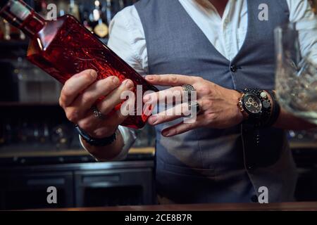 Giovane barista professionista dal taglio corto che tiene una bottiglia rossa durante la preparazione un cocktail al bar Foto Stock