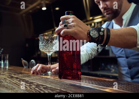 Giovane barista professionista corto che tiene una bottiglia rossa mentre si prepara un cocktail al bar Foto Stock
