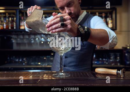 Barista professionista con bearded mirato che versa cubetti di ghiaccio in un cocktail in un bicchiere mentre si lavora in un bar Foto Stock