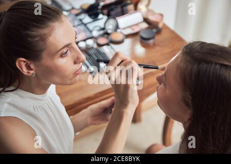 Crop messo a fuoco trucco artista applicando eyeliner nero su bello giovane le palpebre del cliente femminile mentre lavorano in studio contemporaneo Foto Stock