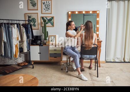 Positivo giovane artista di trucco femminile facendo i clienti faccia trucco usando spazzola vicino a un grande specchio accogliente con luci di bellezza contemporanea salone Foto Stock