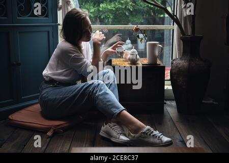 Vista laterale di raccolto calma giovane signora in abito casual versare il tè in una tazza di porcellana durante la cerimonia in un accogliente caffè Foto Stock