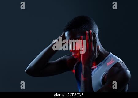 Vista laterale di un brutale atleta africano-americano in abbigliamento sportivo tenere le mani sulla testa e guardare lontano con la sfida mentre in uno studio buio con luci al neon rosse Foto Stock