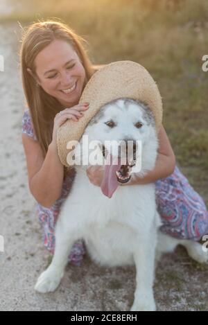 Donna felice in abito che gioca lungo la strada sabbiosa con l'Alaskan Malamute mentre vi godete il fine settimana durante il tramonto in campagna Foto Stock