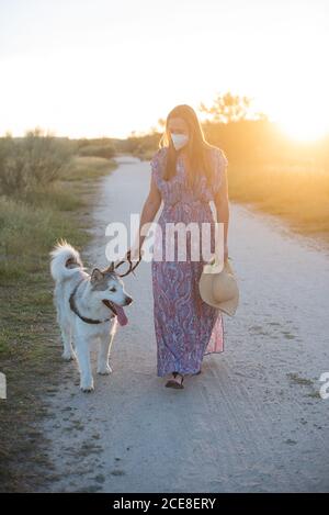 Donna felice in abito a piedi lungo la strada sabbiosa con Alaskan Malamute mentre vi godete il fine settimana durante il tramonto in campagna Foto Stock