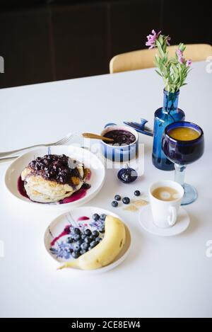 Da sopra deliziosi pancake fatti in casa con marmellata di mirtilli serviti con tazza di caffè, frutti di bosco freschi e banana sul tavolo impostazione per il brunch in casa in una cucina accogliente Foto Stock