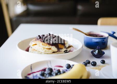 Da sopra deliziosi pancake fatti in casa con marmellata di mirtilli serviti con tazza di caffè, frutti di bosco freschi e banana sul tavolo impostazione per il brunch in casa in una cucina accogliente Foto Stock