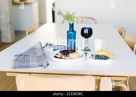 Da sopra deliziosi pancake fatti in casa con marmellata di mirtilli serviti con tazza di caffè, frutti di bosco freschi e banana sul tavolo impostazione per il brunch in casa in una cucina accogliente Foto Stock