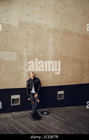 Corpo pieno sicuro giovane calvo maschio in casual giacca nera e jeans che guardano via mentre si levano in piedi contro la costruzione di cemento sopra via Foto Stock