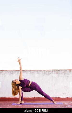 Vista laterale del reggiseno flessibile e delle leggings che si esercitano yoga in posizione laterale estesa sul tappetino sulla terrazza Foto Stock