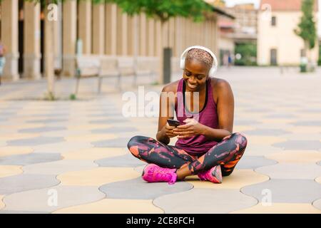 Atleta afroamericana positiva in abiti sportivi e cuffie seduta appoggiata in avanti con gambe incrociate sul terrapieno vicino all'edificio navigazione in città sullo smartphone durante l'ascolto di musica Foto Stock