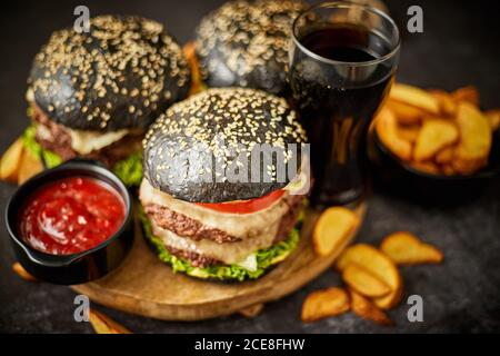 Deliziosi hamburger neri fatti in casa serviti su un tagliere di legno con ketchup e zeppe di patate Foto Stock