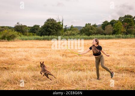 Vista laterale della donna felice in abiti casual che tengono il giocattolo mentre si gioca con adorabile cane grande sul prato in autunno sotto cielo nuvoloso Foto Stock