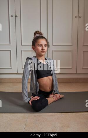 Dall'alto sottile ragazza adolescente seduta su tappetino facendo splits pratica ginnastica a casa guardando via Foto Stock