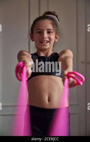 Felice adolescente ragazza in nero sportswear che si riscalda con rosa fascia elastica che sorride alla telecamera Foto Stock