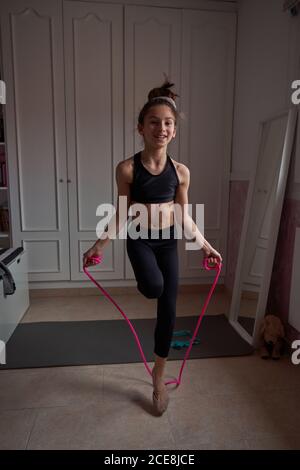 Corpo pieno di teen slender ginnast in nero sport salto corda mentre si allenano nella propria stanza a casa guardando fotocamera Foto Stock