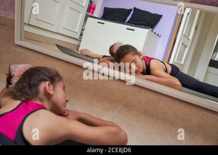 Sottile ragazza adolescente seduta su tappetino facendo split con chiuso occhi che praticano la ginnastica di fronte allo specchio Foto Stock