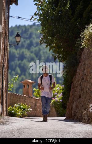 Lunghezza completa di turista maschile elegante bearded in outfit alla moda e occhiali da sole con zaino che cammina sul ponte nella città vecchia situato in verdi colline nel giorno d'estate Foto Stock