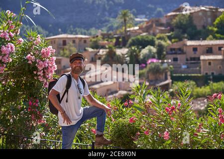 Vista laterale del turista hipster maschile in elegante vestito con zaino in piedi vicino recinto con cespugli fioriti e godendo pittoresco scenario della città vecchia situato tra verdi colline in estate giorno Foto Stock