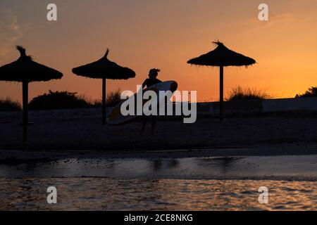 Silhouette di donna anonima con surf a piedi lungo la spiaggia durante il tramonto incredibile in estate