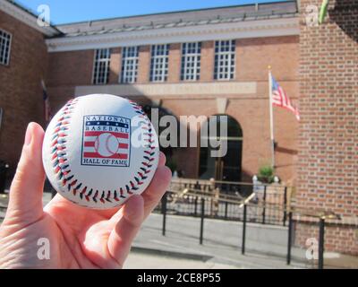 Cooperstown, NY / USA - 3 giugno 2019: Un baseball con il logo della National Baseball Hall of Fame di fronte al Hall Museum Foto Stock