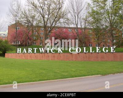 Oneonta, NY / USA - 11 maggio 2019: Cartellonistica Hartwick College che mostra il campus con alberi graziosi, erba verde e grande sala di residenza in mattoni sullo sfondo. Foto Stock