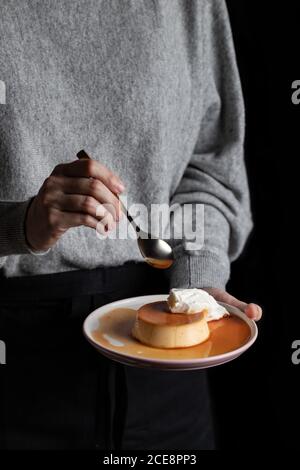 donna irriconoscibile che mangia deliziosa crema di caramello messo su piatto con panna montata su sfondo nero Foto Stock