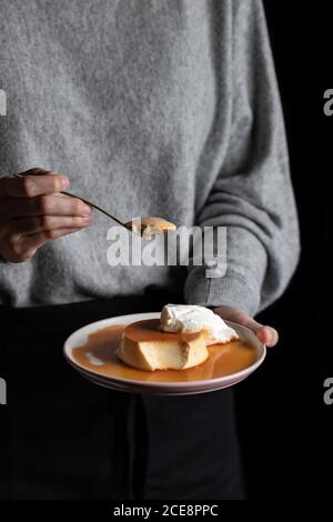 donna irriconoscibile che mangia deliziosa crema di caramello messo su piatto con panna montata su sfondo nero Foto Stock