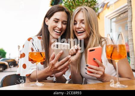 Immagine di due donne adulte sorridenti e che usano i telefoni cellulari mentre si bevono dei cocktail al tavolo in un caffè all'aperto Foto Stock