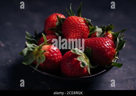 Primo piano di deliziose fragole mature succose con foglie verdi arrangiate in ceramica nera su fondo scuro Foto Stock